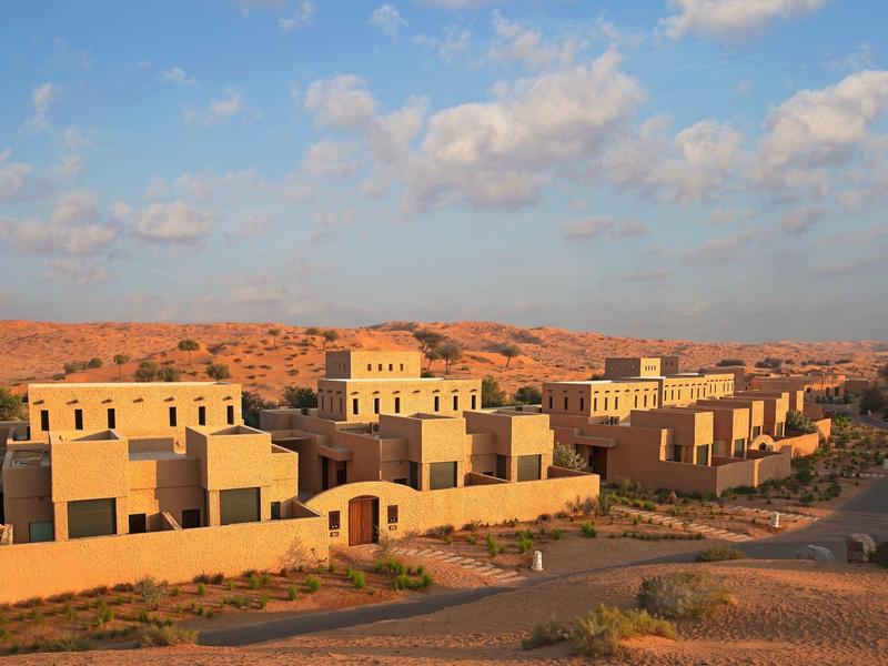 Wohnhäuser in heller Sandsteinfarbe liegen in einer trockenen Wüstenlandschaft unter blauem Himmel.