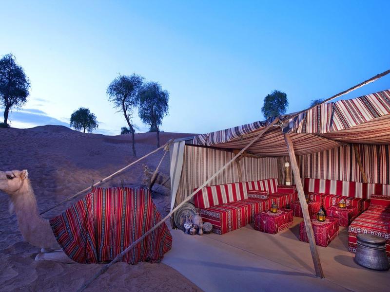 Rotes Kamel mit Sattel vor großem, gestreiftem Zelt in Wüstenlandschaft bei Abendlicht.