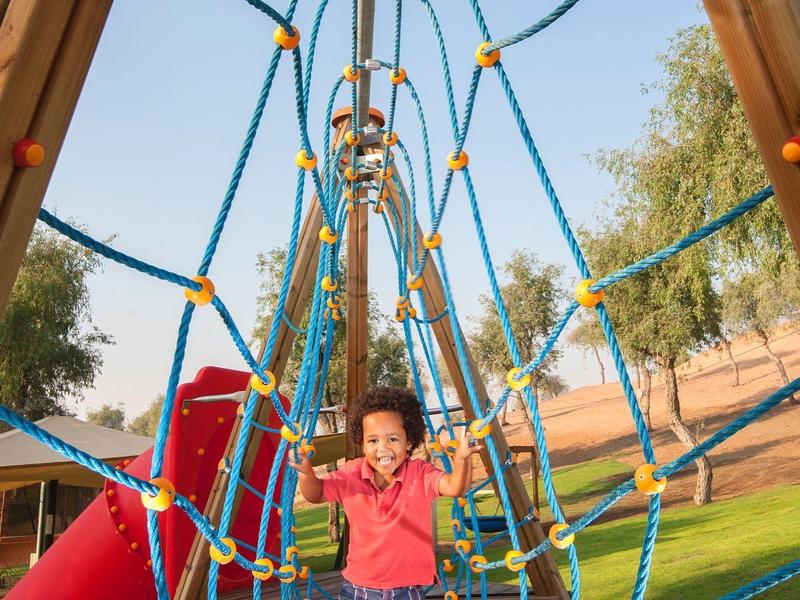 Kind spielt auf blau-rotem Seilnetz in sonnigem Park mit Rutschen und Bäumen im Hintergrund.