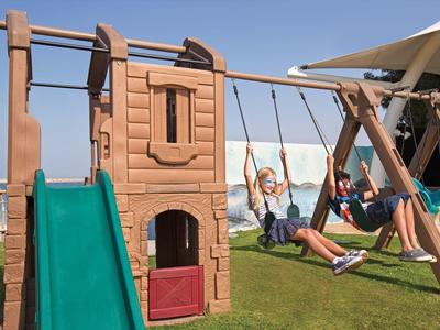 Kinder spielen auf einem Holzspielplatz mit Rutsche und Schaukeln auf einer grünen Wiese am Meer.