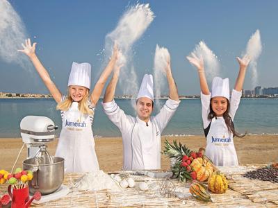 Drei Personen mit Kochmützen posieren am Strand mit Meeresblick und frischen Früchten.