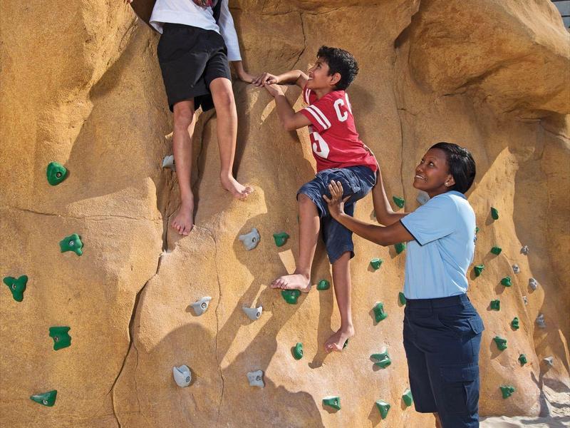 Drei Personen klettern an einer künstlichen Felswand im Sand unter blauem Himmel.