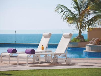 Deux chaises longues blanches avec coussins violets au bord d'une piscine avec vue sur la mer et un palmier.