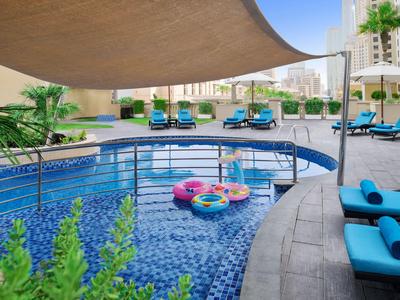 Nowoczesny basen hotelowy z niebieskimi leżakami, żaglem przeciwsłonecznym i kolorowymi dmuchanymi kołami w słoneczną pogodę.