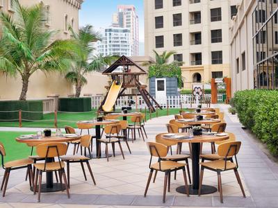 Zewnętrzny teren hotelu z stolikami i krzesłami na tarasie obok małego placu zabaw.