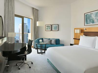 Chambre d'hôtel moderne avec grand lit, bureau, canapé bleu et balcon avec vue sur la ville.