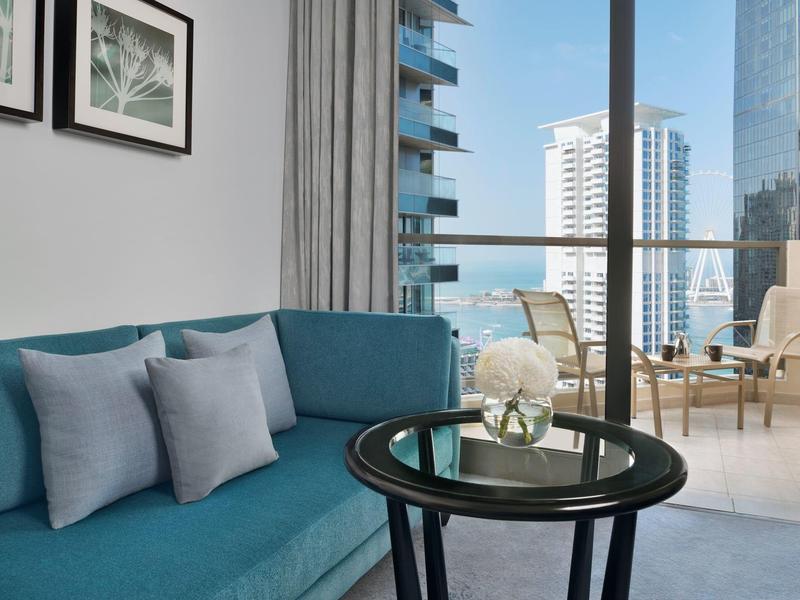 Salon moderne avec canapé bleu, coussins, table en verre, chaises de balcon et vue sur la ville.