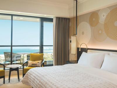 Chambre d'hôtel moderne avec grand lit et vue sur la mer à travers une façade vitrée.