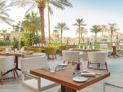 Outdoor restaurant with wooden tables and white chairs beside palm trees and a pool.