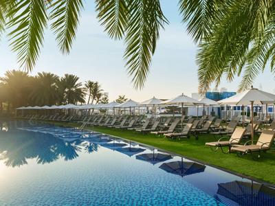 Großer Hotelpool mit Liegestühlen und Sonnenschirmen, umgeben von Palmen bei Sonnenuntergang.