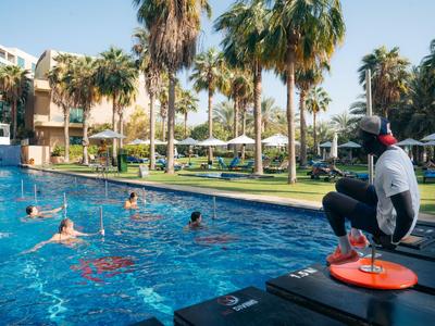 Osoba obserwuje pływaków na basenie na zewnątrz obok palm i budynków hotelu.