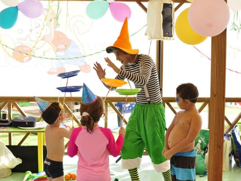 Clown in buntem Kostüm unterhält drei Kinder mit Hüten und Luftballons.