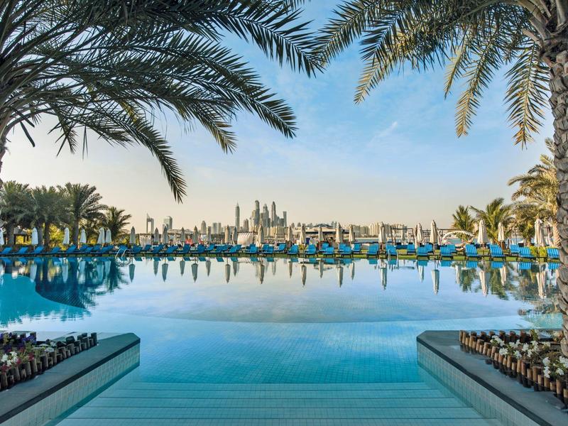 Großer Pool mit Palmen und Blick auf die Skyline einer Stadt bei klarem Himmel
