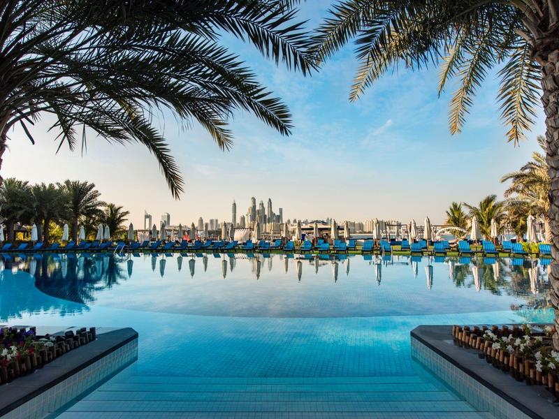 Blick auf einen Pool mit Palmen und der Skyline einer Stadt im Hintergrund bei klarem Himmel.