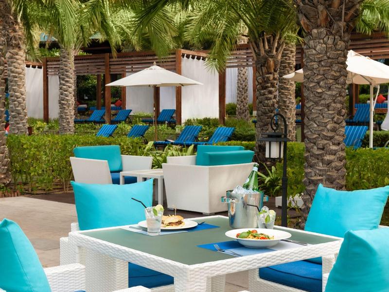 Moderner Außenbereich mit blauen Sitzkissen, Tischen und Sonnenschirmen in tropischer Umgebung.