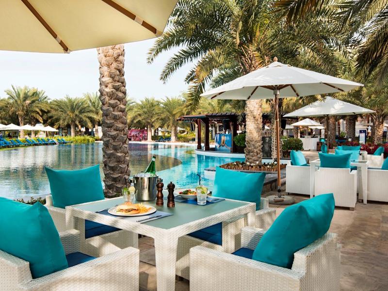 Outdoor-Essbereich mit weißen Möbeln und türkisfarbenen Kissen neben einem Pool mit Palmen.