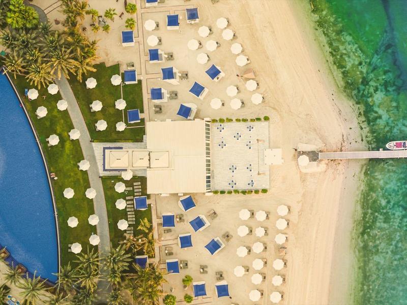 Vogelperspektive eines Resorts mit Pool, Strandliegen und einem Steg im türkisblauen Meer.