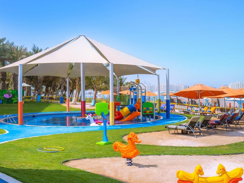 Kinderspielplatz mit Wasserspielbereich, Sonnenschirmen und Liegestühlen in einem Hotelgarten