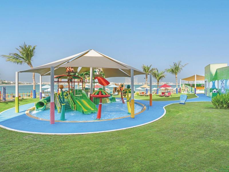 Bunter Kinderspielplatz mit Rutschen und Klettergerüsten neben einem grünen Rasen und Palmen unter blauem Himmel.