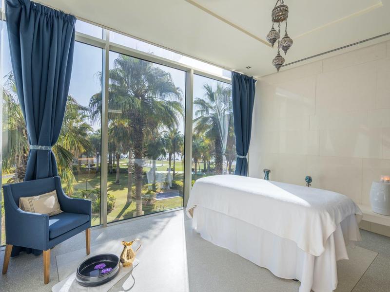 Behandlungsraum mit Massageliege, blauem Sessel und großen Fenstern mit Blick auf Palmen.