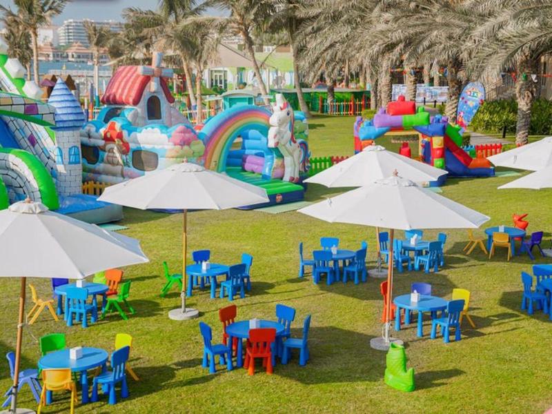 Bunte Kindersitze mit weißen Sonnenschirmen auf grüner Wiese vor großen, farbigen Hüpfburgen.