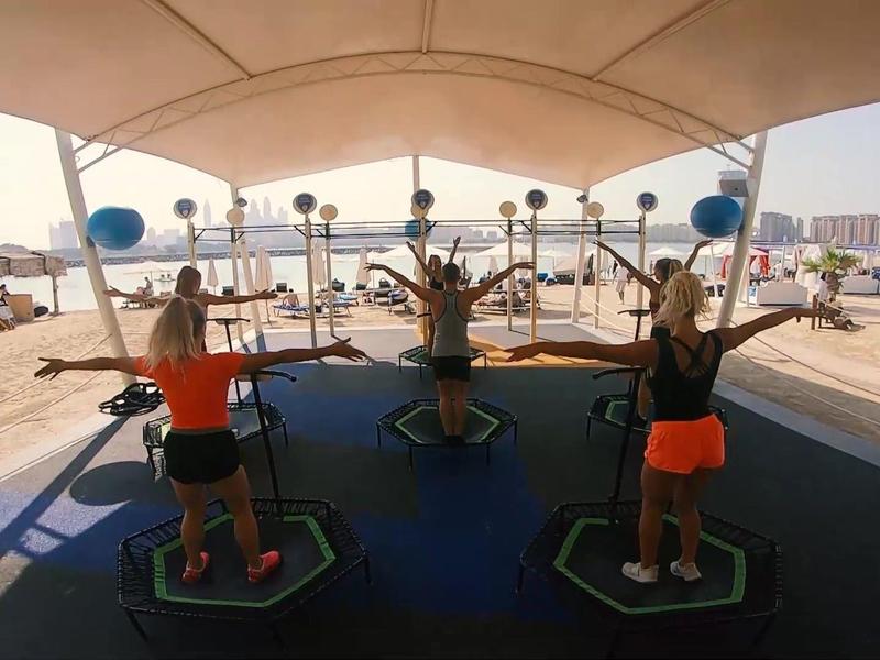 Drei Frauen springen auf einzelnen Trampolinen unter einem Sonnendach am Strand.
