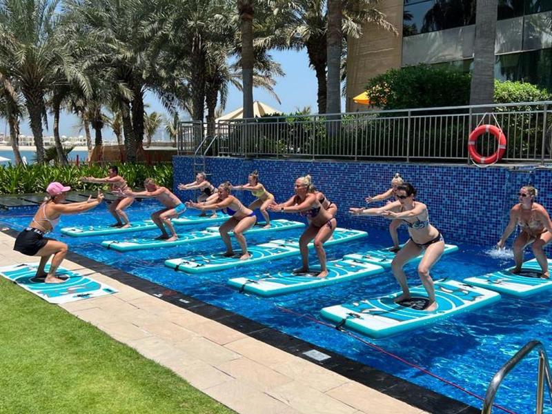 Menschen machen Aqua-Fitness im Pool, umgeben von Palmen und blauer Mauer, bei sonnigem Wetter.