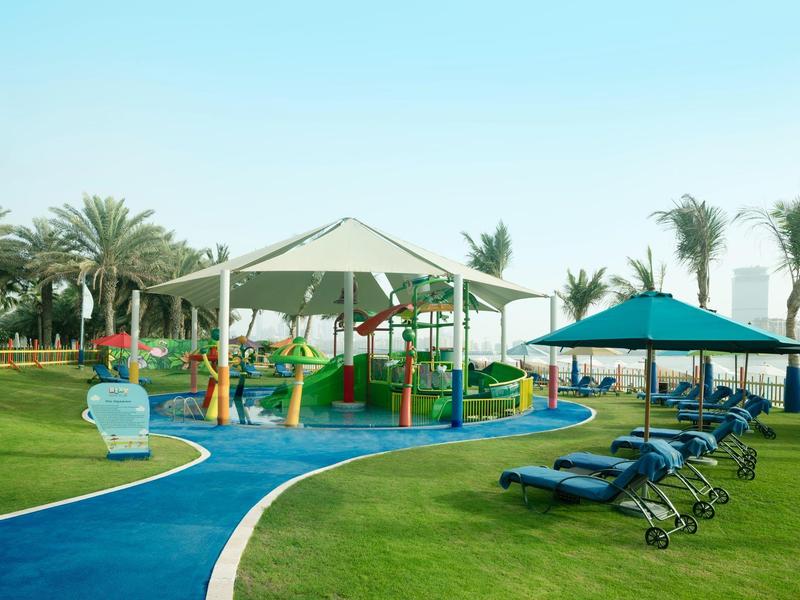 Spielplatz mit Pavillon und Rutschen neben Liegestühlen auf einer grünen Wiese und Palmen.