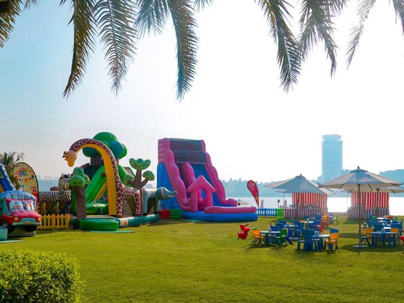 Grüner Garten mit bunten aufblasbaren Rutschen, Palmen und Sonnenschirmen im Hintergrund.