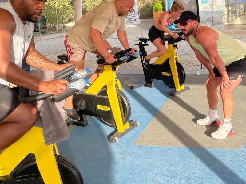 Drei Männer trainieren intensiv auf gelben Fitness-Fahrrädern unter einem Sonnenschutz.
