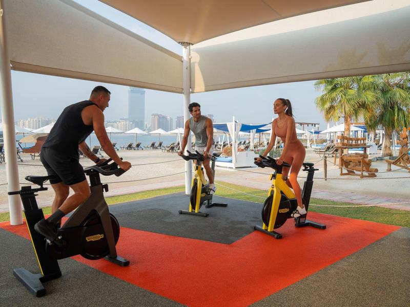 Drei Personen trainieren auf Indoor-Fahrrädern unter einem offenen Zelt in Strandnähe.