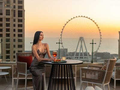 Frau im eleganten Kleid sitzt auf Balkon mit Tischen, Stühlen und großem Riesenrad im Sonnenuntergang.