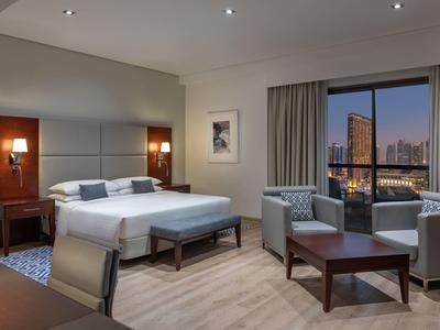 Camera d'hotel moderna con letto matrimoniale accogliente, zona salotto e vista sulla città di notte.