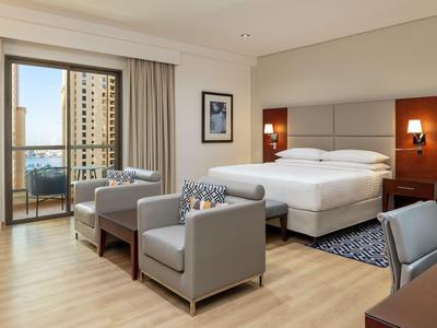 Camera d'hotel luminosa con letto matrimoniale, poltrone, scrivania e balcone con vista sulla città.