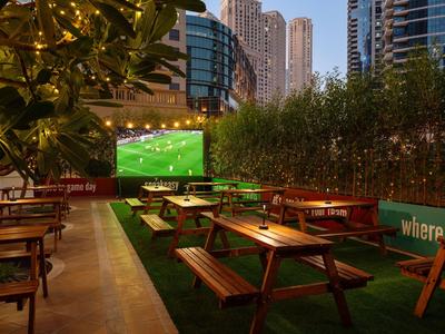 Outdoor terrace with wooden tables and benches facing a large screen showing a soccer match.