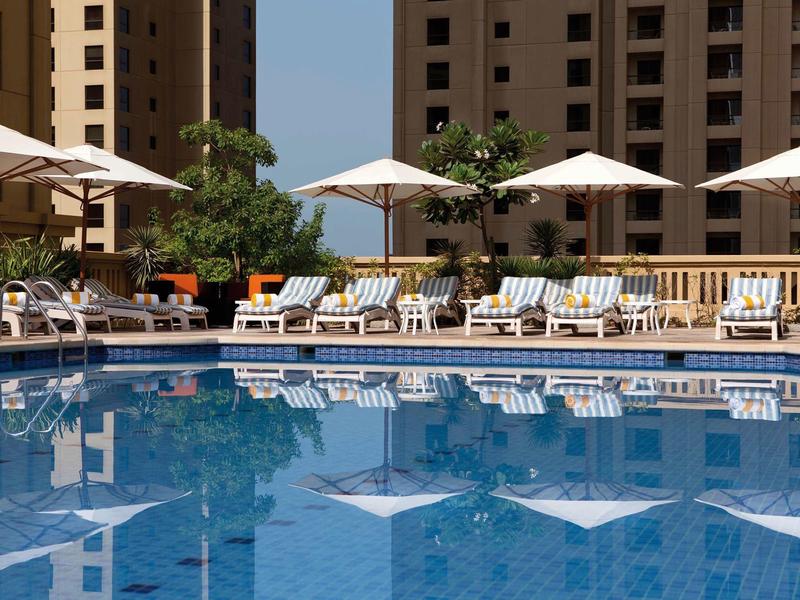 Duży basen hotelowy z leżakami i parasolami przed wysokimi budynkami.