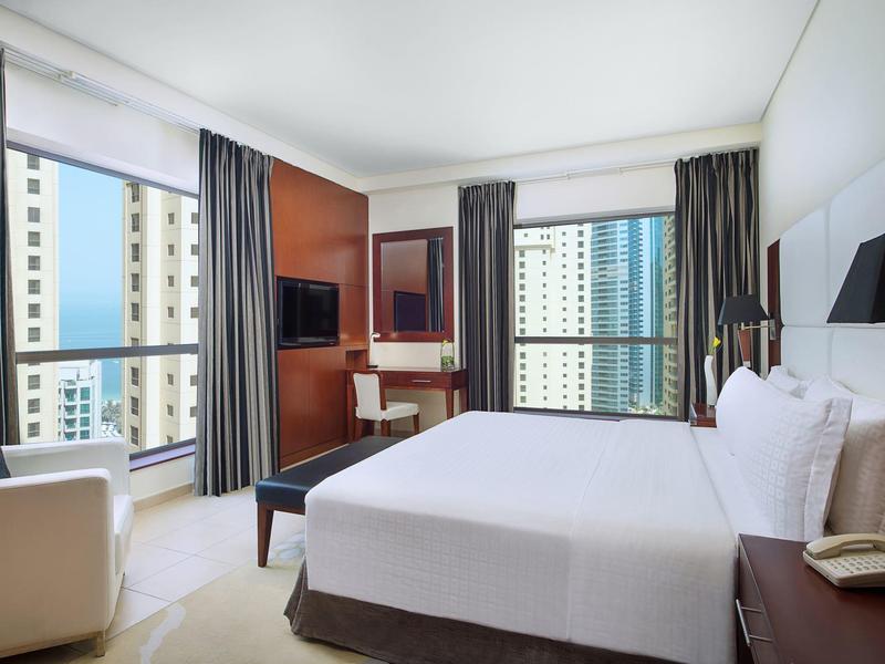 Jasny pokój hotelowy z podwójnym łóżkiem, biurkiem, fotelami i dużymi oknami z widokiem na miasto.