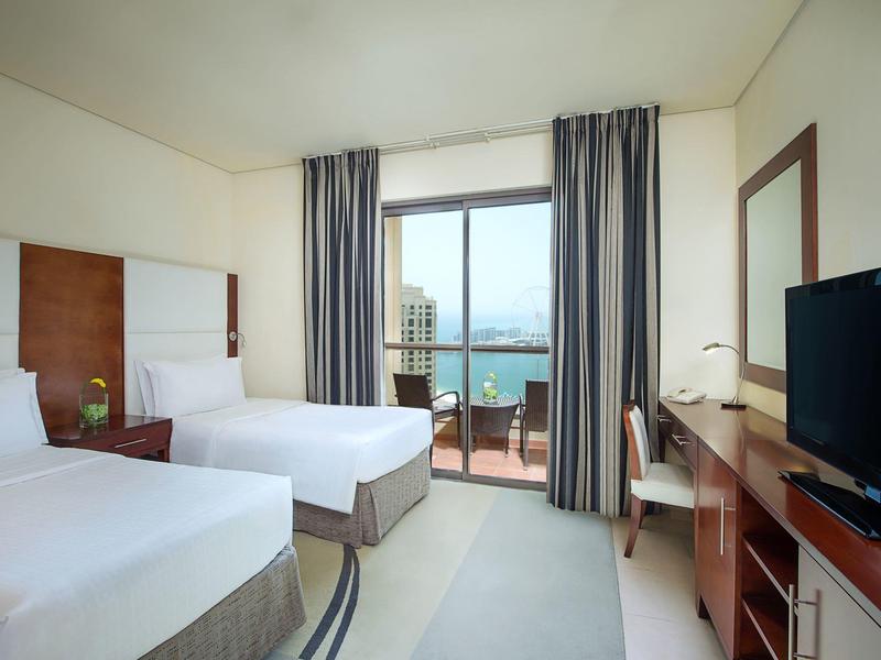 Pokój hotelowy z dwoma łóżkami, telewizorem, biurkiem i balkonem z widokiem na morze.