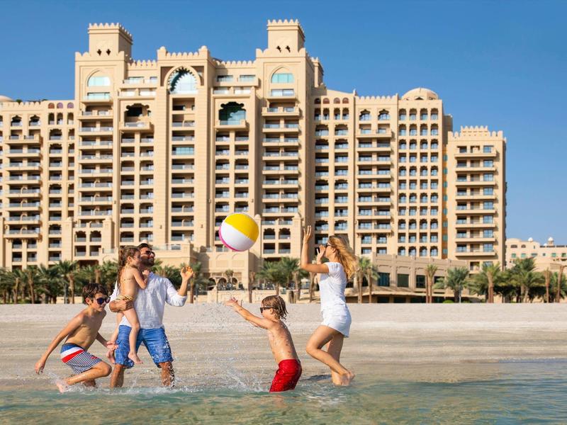 Familie spielt mit einem bunten Ball am Strand vor einem großen Hotel mit Sandtürmen.