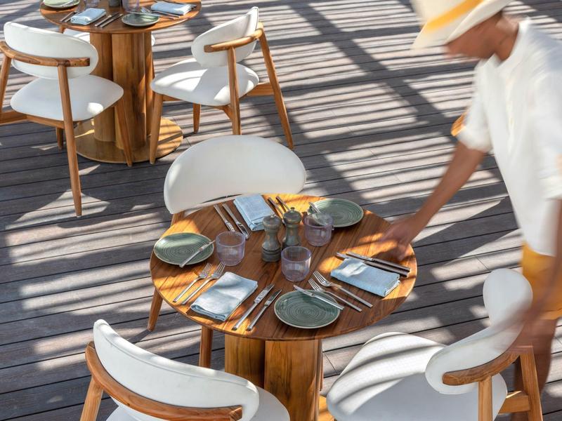 Ein gedeckter Tisch mit Stühlen auf einer Terrasse mit Schatten, eine Person in Bewegung.