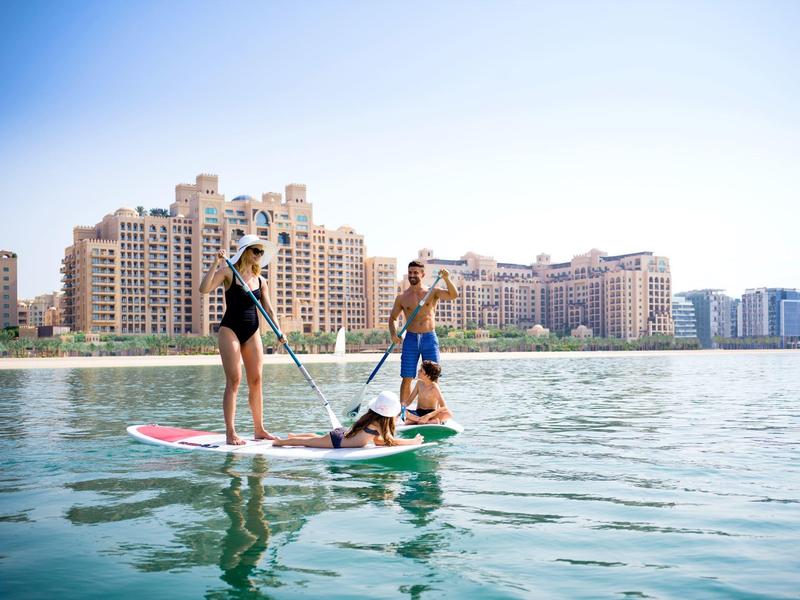 Drei Personen paddeln auf einem großen Stand-Up-Paddleboard in der Nähe von Gebäuden am Wasser.