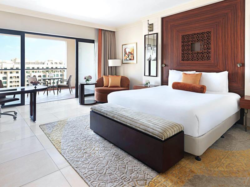 Helles Hotelzimmer mit großem Bett, Schreibtisch, Sesseln und Balkon mit Stadtblick.