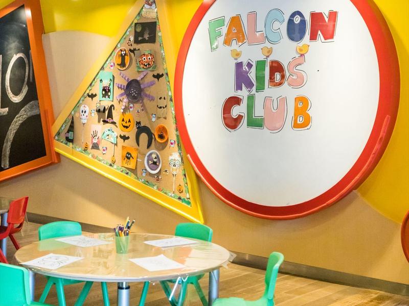 Bunter Kinderclub mit runden Tischen und türkisfarbenen Stühlen vor gelber Wand.