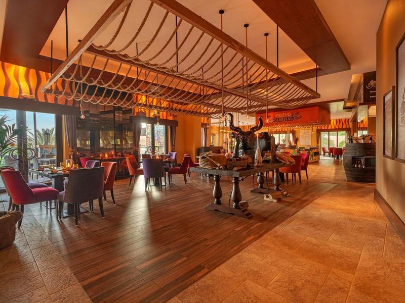 Ein warm beleuchtetes Restaurant mit Holzboden, rot gepolsterten Stühlen und großen Fenstern.