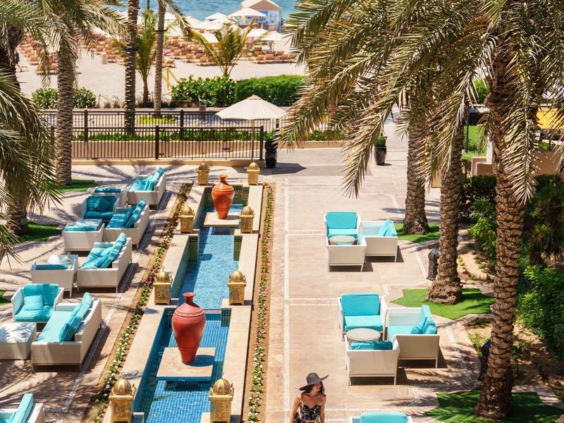 Hotelpoolbereich mit blauen Liegen, Palmen und Blick auf Meer und Strand im Hintergrund.