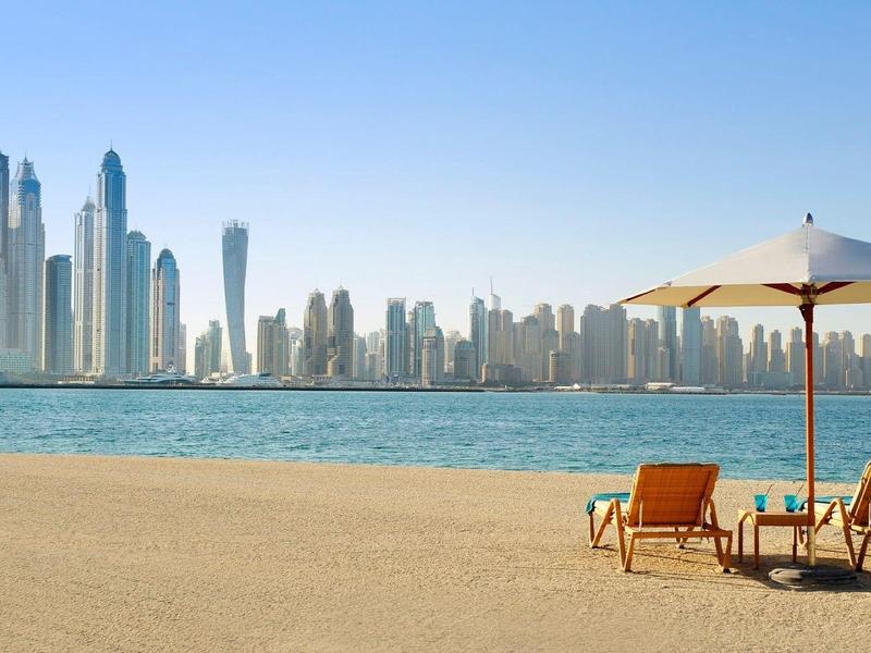 Leerer Strand mit zwei Sonnenschirmen und Liegestühlen vor moderner Skyline am Wasser.