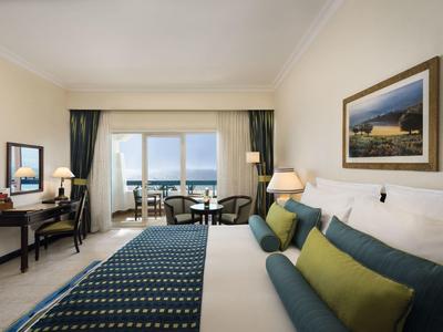 Helles Hotelzimmer mit Doppelbett, Sitzbereich und Balkon mit Meerblick.