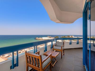 Balkon mit Holzstühlen, blauem Geländer und weitem Blick auf Meer und Strand unter klarem Himmel.