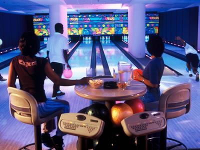 Zwei Frauen sitzen an einem Tisch in einer Bowlingbahn mit Bahnen und Spielern im Hintergrund.