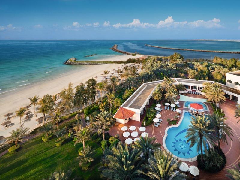 Hotel mit Pool, Palmen und Strand am blauen Meer unter blauem Himmel mit wenigen Wolken.